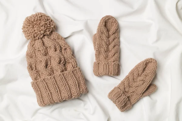 Brown winter hat and mittens on white bed sheet. Top view.