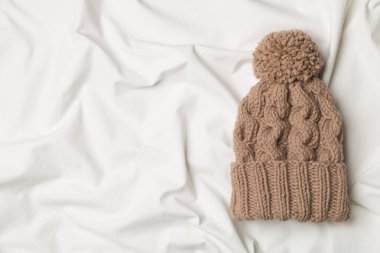 Brown winter hat with pompon on white bed sheet. Top view.