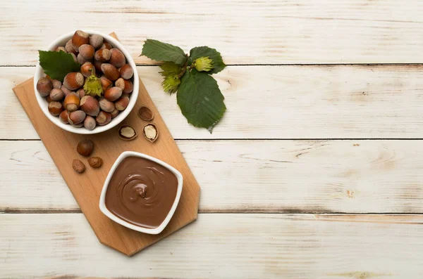 stock image Hazelnut cream with nuts and green leaves on wooden background, top view.