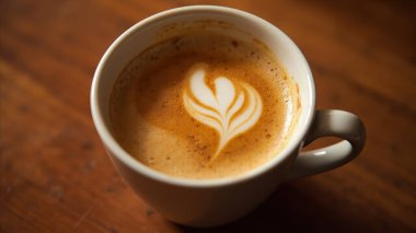 Beyaz bardakta nefis latte sanatı sergileyen yakın çekim çekiminin sıcaklığının tadını çıkar. Kremalı köpük narin bir çiçek deseni ve kalp şekli gösteriyor. Sanatsal becerinin bir kanıtı. Zengin ahşap bir masada dinleniyor, bu taze ve gurme kaftan