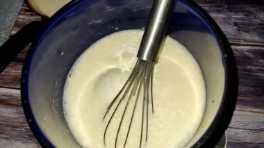 Adding wheat flour to beaten eggs and milk in a blue saucepan.  Preparing pancake dough