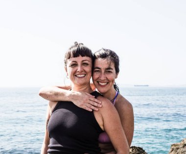 İki gülümseyen arkadaş okyanus kıyısında sıcak bir kucaklaşmayı paylaşıyor, kumsalda mutluluk ve dostlukla dolu güneşli bir günün tadını çıkarıyorlar..