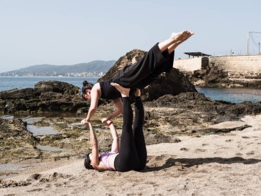 İki kadın kumsalda akro yoga yapıyor. Kum ve kayaların üzerinde zarif bir şekilde dengede duruyorlar. Manzaralı bir okyanus zeminiyle. Görüntü, açık hava ortamında uyum, güç ve sükunet sağlar..
