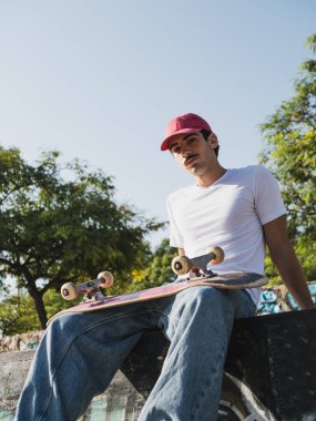 Kırmızı şapkalı kaykaycı güneşli bir kaykay parkında öylesine oturuyor..