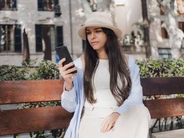 Yazlık şapkalı kadın dışarıdaki ahşap bir bankta akıllı telefon kullanıyor.