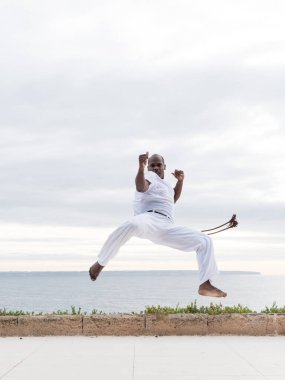 Capoeira dövüş sanatları ustası, açık denizde beyaz giyerken yüksek bir tekme atıyor.
