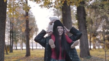 Anne ve küçük çocuk sonbahar parkında. Mutlu bir aile, anneyle yaşamak, neşeli bir çocuk, annesinin omuzlarında oturmak, doğa yürüyüşü.
