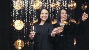 Two young beautiful smiling girls in black jacket. Sexy carefree women posing near silver shiny tinsel wall in studio. Holiday with sparklers and champagne. Happy day