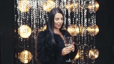 Young beautiful smiling brunette female in black jacket. Sexy carefree woman posing near silver shiny tinsel wall in studio. Fashionable model with makeup holding glass of champagne. Celebration