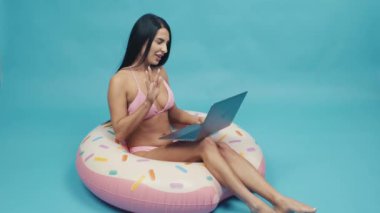 Beautiful woman smiling siting on rubber ring and talking on a laptop on blue background isolated. Happy trip concept