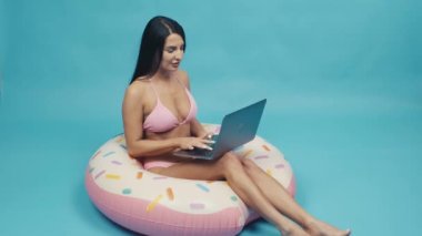 Beautiful woman smiling siting on rubber ring and talking on a laptop on blue background isolated. Happy trip concept