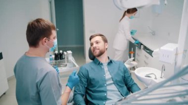 A male doctor discusses a dental treatment plan with a patient. Dentistry. Dental clinic. In the background, a female doctor is preparing a patient