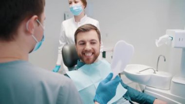 Professional male dentist show beautiful teeth in mirror to man visitor. Positive male patient of dental clinic enjoying amazing smile after treatment