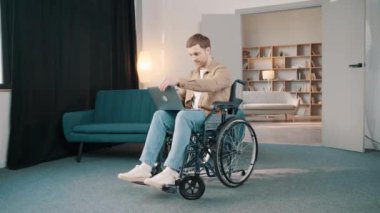 Evdeki oturma odasında dizüstü bilgisayar kullanarak tekerlekli sandalyedeki neşeli, engelli beyaz adam.