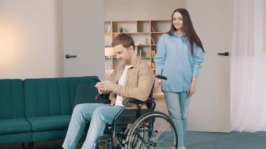Beyaz kadın, genç kocasını destekliyor. Evde tekerlekli sandalyede oturuyor. Çekici bir kadın, erkek hastasıyla ilgilenir. Oturma odasında sağlık için fizik tedavi uygular.