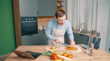 Önlük giyen neşeli adam modern mutfakta pizza hazırlıyor. Erkek domatesleri kesip sosla hamurun üzerine koyar.