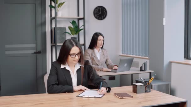 眼鏡のビジネス ガールが事務所でメモを取ってる 女子社員が後ろに座ってる — ストック動画