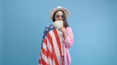 Beyaz şapkalı zengin tatlı kız Amerikan bayrağı ve mavi arka planda gözlüklü kameraya bakarken nakit gösteriyor. Vatandaş ve vatansever kızın portresi. İçeride yavaş çekimde para ile poz veriyor. Yavaş çekim