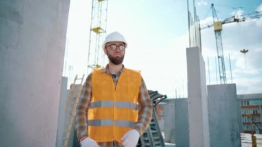 Gülümseyen, kendine güvenen, üniformalı, kask ve gözlük takan, inşaat alanının önünde kameraya bakan genç bir işçinin portresi.