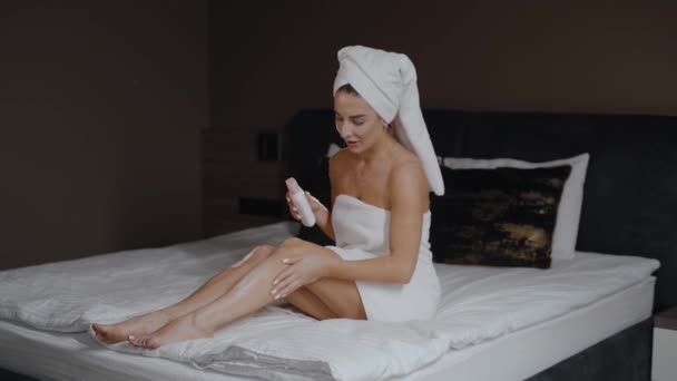 一个头戴毛巾的快乐女人正坐在床上 把奶油放在腿上 享受着放松的时光 美丽的高加索女人在家里的卧室里洗澡后躺在床上休息 — 图库视频影像
