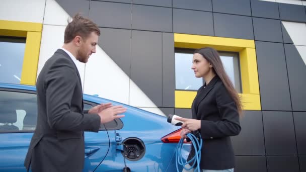 ディーラーのビジネスマンは 充電ステーションで電気自動車を充電する方法を女の子に示します 女性はパワーコードを屋外の電気自動車に接続します スローモーション — ストック動画