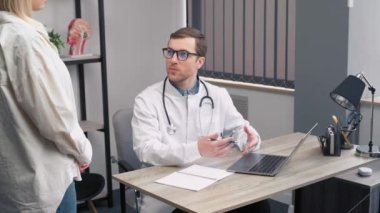 Tıbbi üniformalı ve steteskoplu bir erkek ent doktoru klinikteki masada oturmuş, muayene sırasında kadın hastayla konuşuyor. Doktor bilgileri bir deftere yazar.