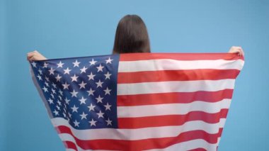 Güzel, çekici, esmer bir kadın Amerikan bayrağını sarıp, Bağımsızlık Günü 'nü kutluyor, insan hakları ve özgürlüklerini kutluyor, stüdyoda mavi renkli arka plan üzerine izole edilmiş pozlar veriyor.