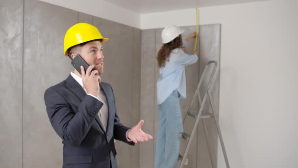 穿着西装的商人 公寓里戴着黄色的硬礼帽 一个在电话里说话的男人 一个背靠梯子的带尺子的女工 — 图库视频影像