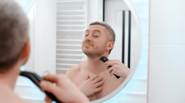 Aynadaki yansımanın arkasındaki erkek aynası. Banyodaki çıplak adam. Sakallı beyaz erkek esmer. Evde tıraş olurken çenesini tıraş etmek için elektrikli tıraş makinesi kullanıyor.