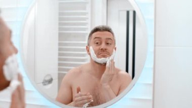 Aynaya bak, tıraş köpüğü, hijyen, güzellik ve cilt bakımını sabah rutiniyle yap. Evdeki tuvalette köpüklü, kozmetik ürünü olan ve kendine özen gösteren mutlu bir erkek.