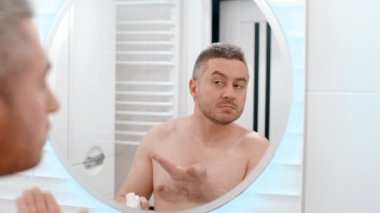 Genç adam banyo aynasına bakarak cildine nemlendirici krem sürüyor. Yakışıklı sakallı adam cilt kremi sürerek sabah rutinini yapıyor.
