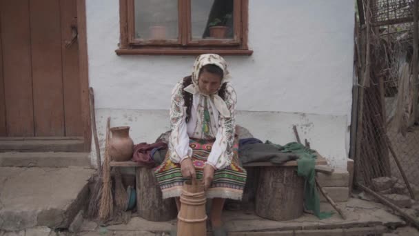 ウクライナのシンボルの老婦人は古い家の近くに座って古い方法で油を抽出します — ストック動画