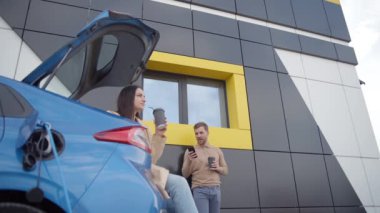 EV şarj istasyonunda kahve içerken gülümseyen ve konuşan mutlu genç çift. Telefonlu bir adam şarj noktasında beklerken kahve içiyor. Sıfır emisyon arabası