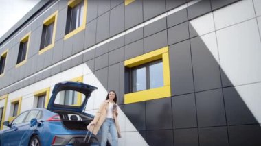 Elinde kahve ve valiziyle beyaz bir kadın elektrikli arabasının yanında yürüyor ve aracın şarj olmasını bekliyor. Kız bavulunu arabaya koyar. Elektrikli arabanın şarj prizi.