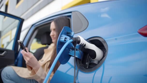Młoda Kobieta Siedzi Elektrycznym Samochodzie Patrzy Smartfon Stacja Ładowania Elektrycznego — Wideo stockowe