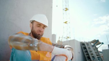 Genç ustabaşı dışarıda oturuyor ve çok çalıştıktan sonra yorgun hissediyor. Kameraya bak. Yansıtıcı yelek ve miğfer giymiş beyaz işçi inşaat alanında. Yavaş çekim