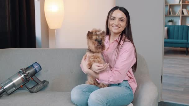 快乐的少女坐在舒适的沙发上 在公寓里和可爱的小宠物狗玩耍 附近的无绳真空吸尘器 — 图库视频影像