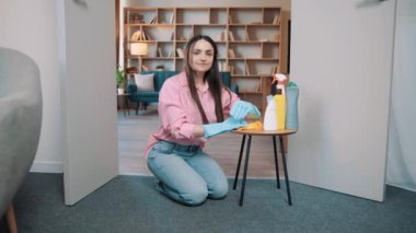 Pembe tişörtlü, büyük lastik eldivenli bir kız evde yere oturup kameraya poz veriyor. Evde deterjanı olan bir kadın.