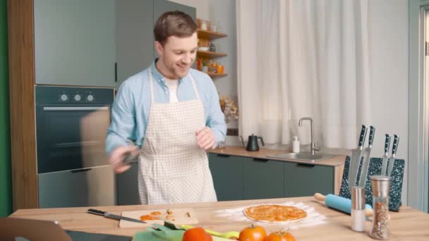 穿着围裙的快乐男人在现代厨房里准备披萨 男人把西红柿和酱汁一起放在面团上 然后用手机拍照 — 图库视频影像