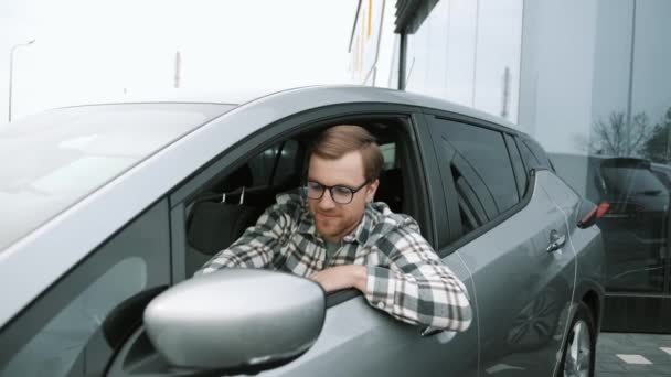 車のディーラーで新しい高級電気自動車を運転幸せな若い男の肖像画 その男は新車を運転しながら幸せの感情を示す 買い手は車を選ぶ — ストック動画