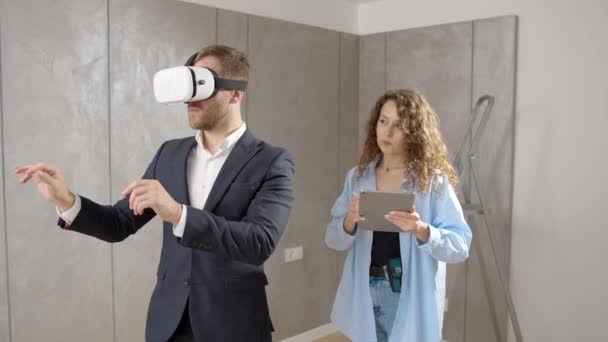 仮想現実 改装の概念を設計するための未来的な技術を使用してVrメガネを身に着けている改装中に新しいアパートに部屋に立つスーツと女性デザイナーの若い男 — ストック動画