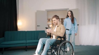 Beyaz kadın, genç kocasını destekliyor. Evde tekerlekli sandalyede oturuyor. Çekici bir kadın, erkek hastasıyla ilgilenir. Oturma odasında sağlık için fizik tedavi uygular.