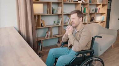 Evde tekerlekli sandalyede yalnız başına oturan genç bir adam pencereye bakıyor. Düşünüyor, düşünüyor, gülümsüyor ve evde kitaplığın yanında poz veriyor.