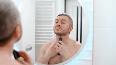 Aynadaki yansımanın arkasındaki erkek aynası. Banyodaki çıplak adam. Sakallı beyaz erkek esmer. Evde tıraş olurken çenesini tıraş etmek için elektrikli tıraş makinesi kullanıyor.