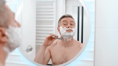 Beyaz sabunlu, köpüklü, sakal tıraşlı jöleli, traş bıçağı olan ve banyoda eğlenen neşeli, komik, beyaz, beyaz, beyaz bir adam.