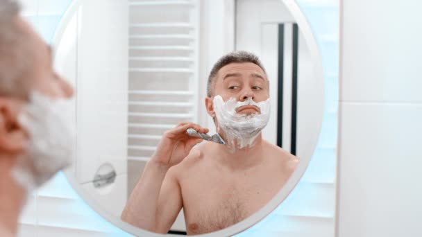 陽気で面白い慎重な白い石鹸のアンヘブン男はひげの表面のシャーブカミソリに発泡ゲルを剃り バスルームで楽しんでいる — ストック動画
