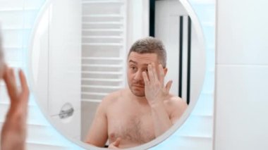Çekici bir genç adam aynaya bakarak kaşlarına nemlendirici krem sürüyor. Yakışıklı sakallı adam sabah cilt bakımını yapıyor.
