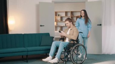 Beyaz kadın, genç kocasını destekliyor. Evde tekerlekli sandalyede oturuyor. Çekici bir kadın, erkek hastasıyla ilgilenir. Oturma odasında sağlık için fizik tedavi uygular.