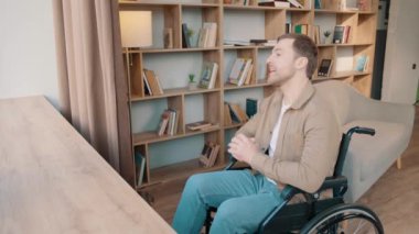 Evde tekerlekli sandalyede yalnız başına oturan genç bir adam pencereye bakıyor. Düşünüyor, düşünüyor, gülümsüyor ve evde kitaplığın yanında poz veriyor.