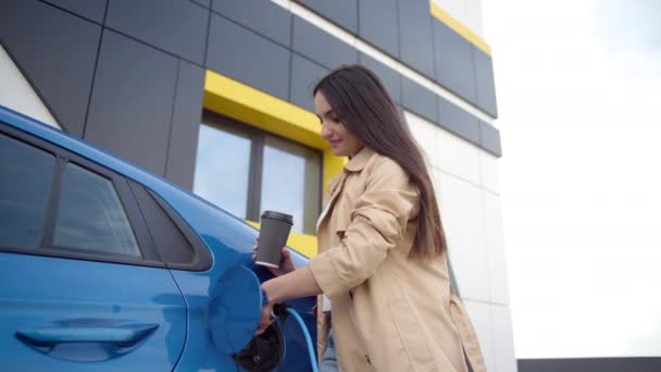 女性は充電器を車の充電ソケットに差し込み 彼女の電気自動車の近くに立っているコーヒーを待ちます 女性は車両が課金されるときに待つ — ストック動画